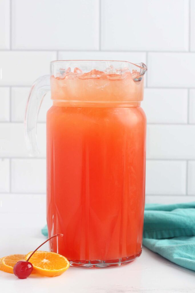 close up of a pitcher sitting on a white background filled with a light redish orange liquid. There is a teal napkin and orange slices with cherries.