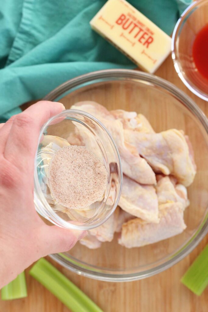 Top down image showing a glass bowl filled with chicken wings being sprinkled with salt
