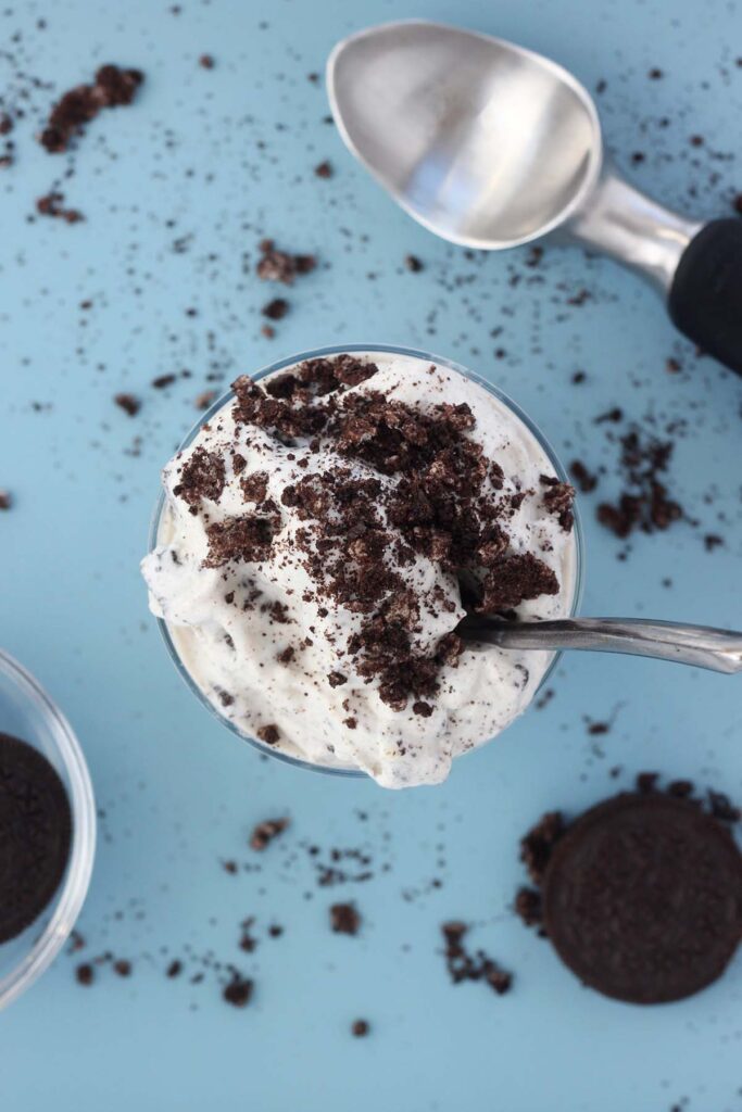 Top down image showing a milkshake with cookie crumbs inside of a glass cup that is sitting on a blue table with cookie crumbs all around