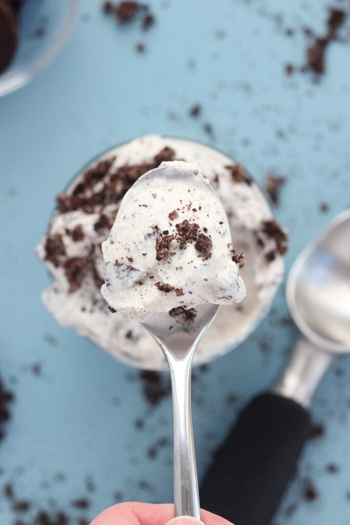 Top down image showing a milkshake with cookie crumbs inside of a glass cup that is sitting on a blue table with cookie crumbs all around. A bite on a spoon is being held above the cup