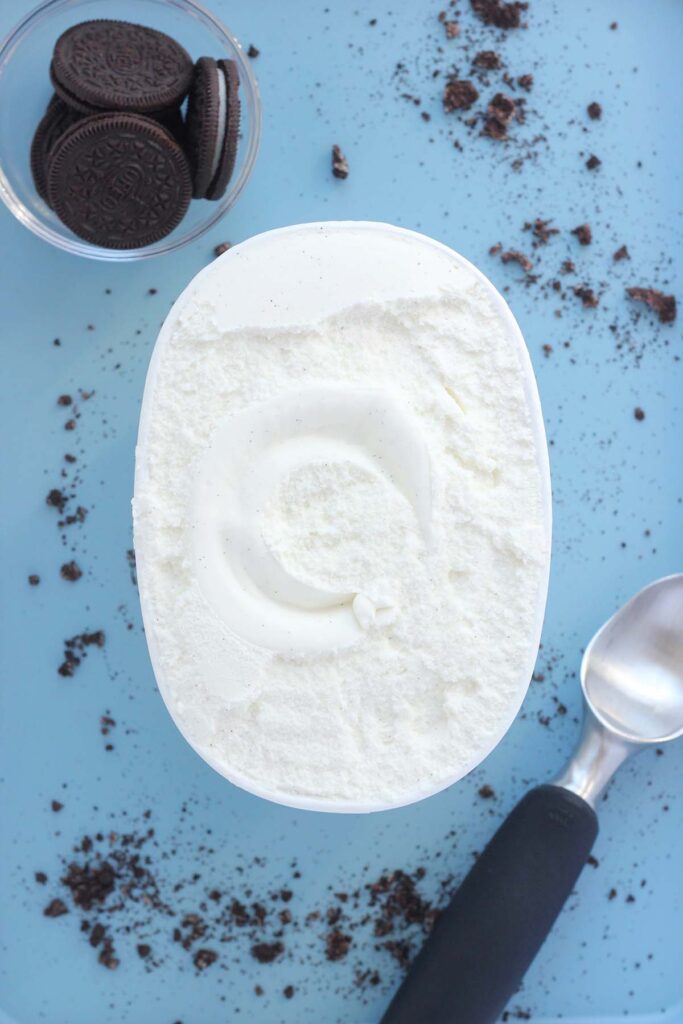 Top down image showing an open container of vanilla ice cream sitting on a blue background that is sprinkled with cookie crumbs and an ice cream scoop
