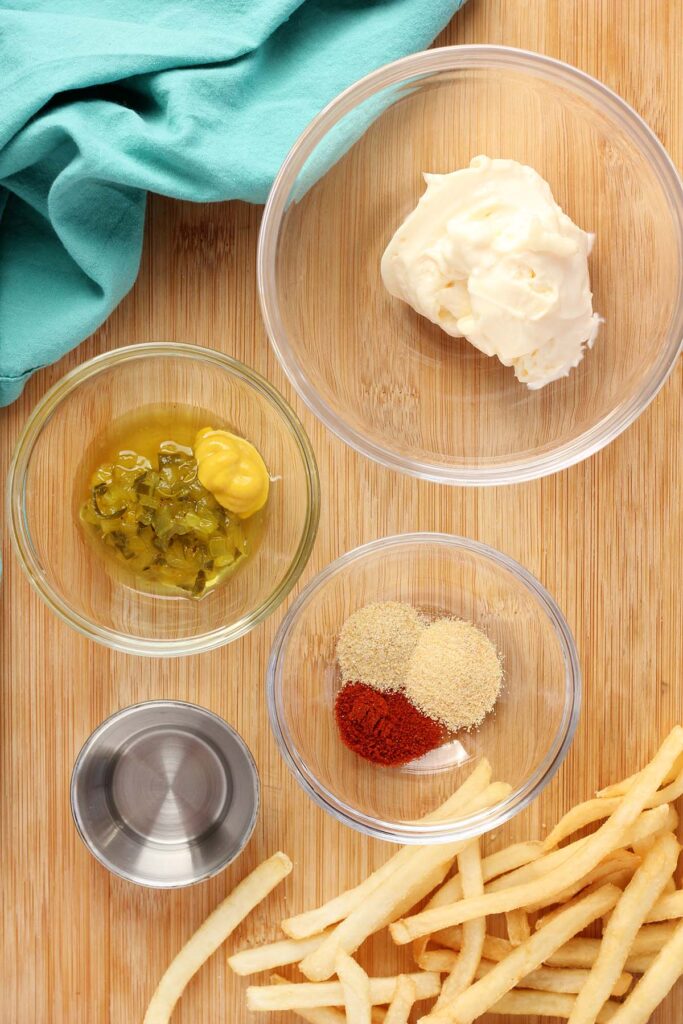 top down image showing a glass bowl with mayo, one with relish and mustard, one with seasonings, and a metal one with vinegar. Fries and a teal napkin are off to the side. 