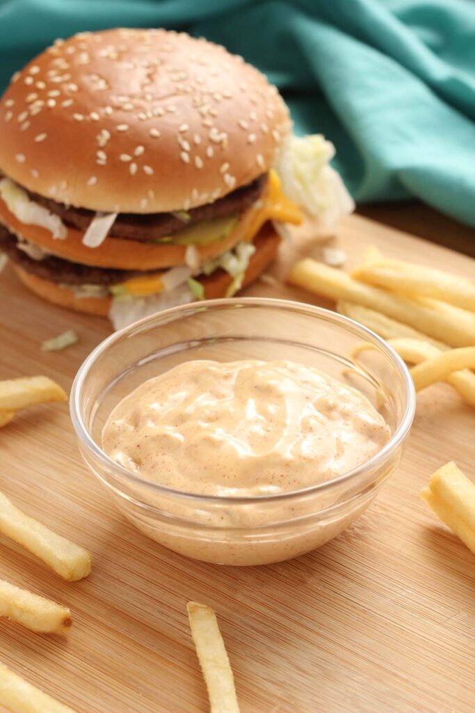 close up of a small glass dish with a sauce. The dish is sitting on a wooden table top with a hamburger off to the side and more fries