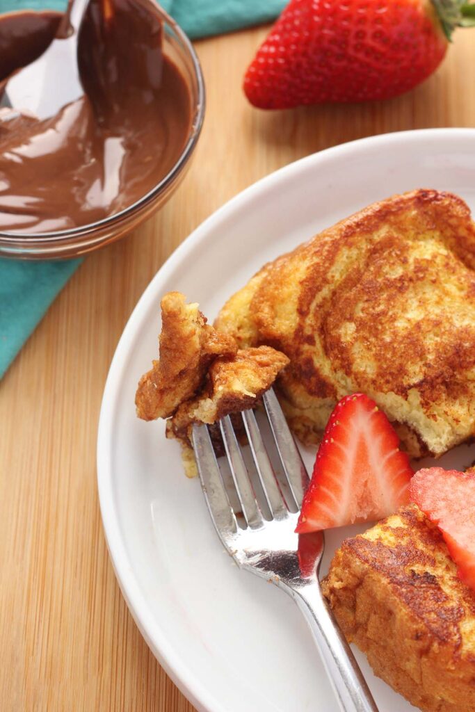 top down image showing french toast topped with strawberries sitting on a white round plate with a bite on a fork
