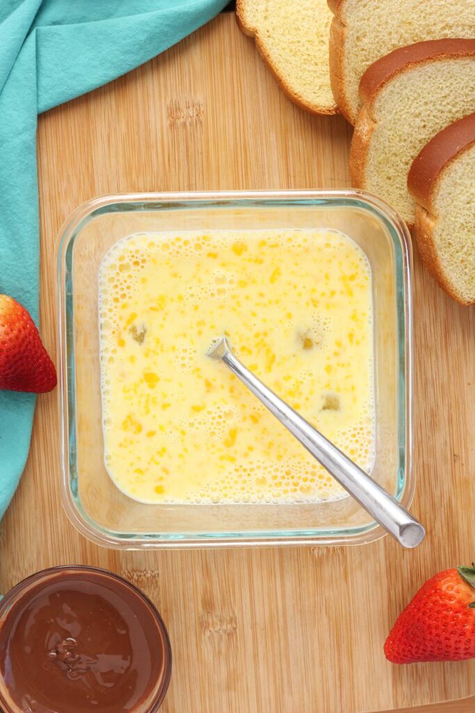 top down image showing a square glass bowl filled with whisked eggs and a fork on a wooden table top with bread, nutella and strawberries off to the sides