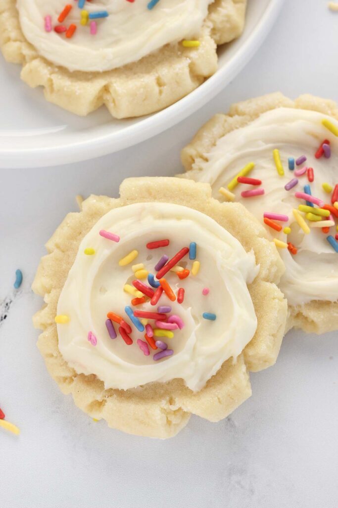 Top down image showing two frosted cookies with rainbow colored sprinkles sitting on a white counter with a plate of cookies off to the side. 