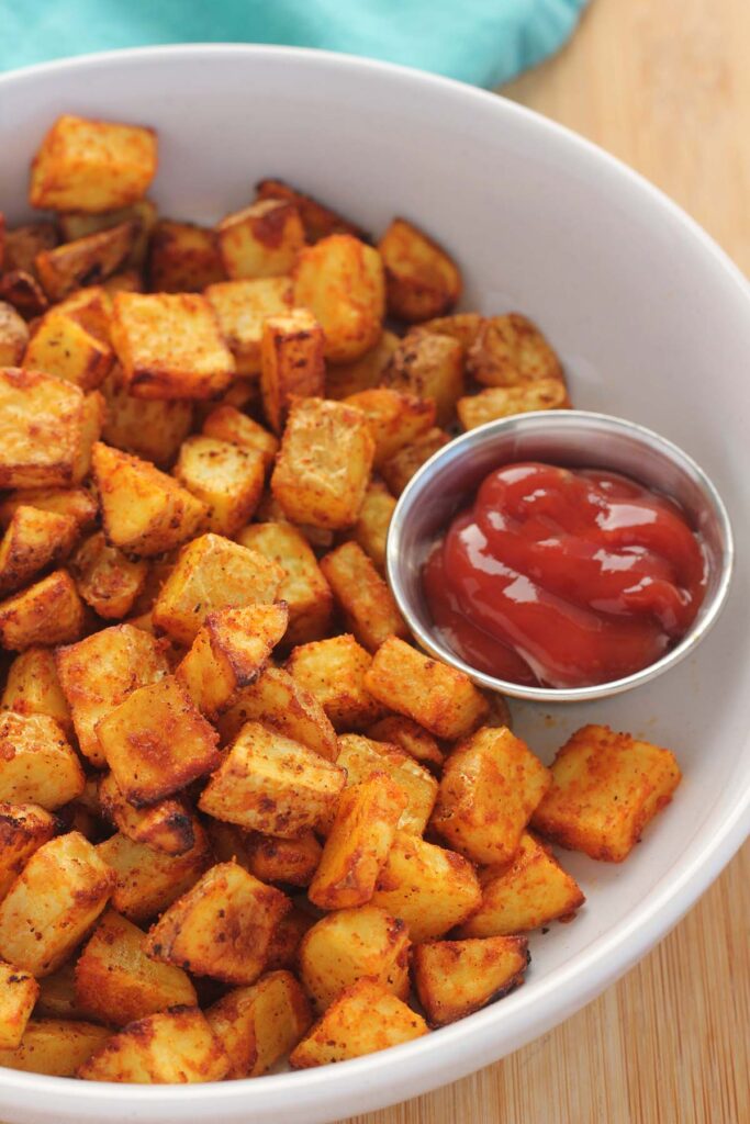Top down image showing cooked potato cubes with a small dish of ketchup in a white bowl 