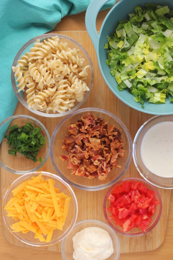 top down image showing a bowl of lettuce, a bowl of pasta, a bowl of bacon, a bowl of onion, bowl of ranch, bowl of cheese, and bowl of tomato