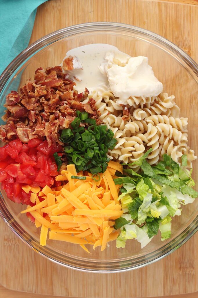 Top down image showing a large glass mixing bowl filled with pasta, lettuce, cheese, tomato, green onion, bacon, ranch and mayo unmixed