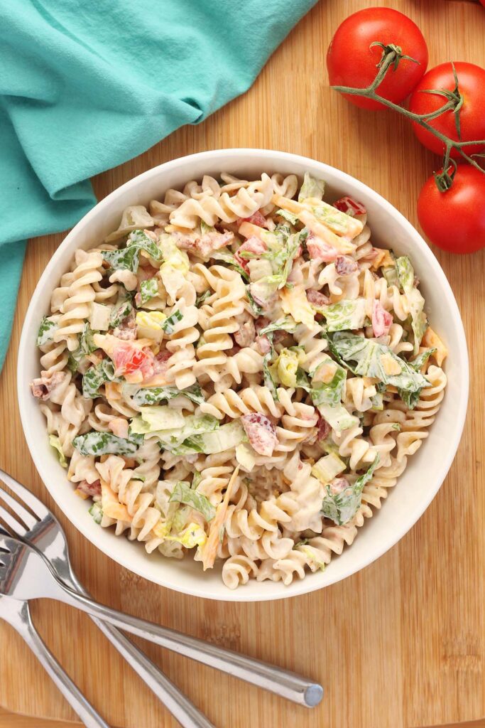 Top down image showing a large white mixing bowl filled with pasta, lettuce, cheese, tomato, green onion, bacon, ranch and mayo mixed