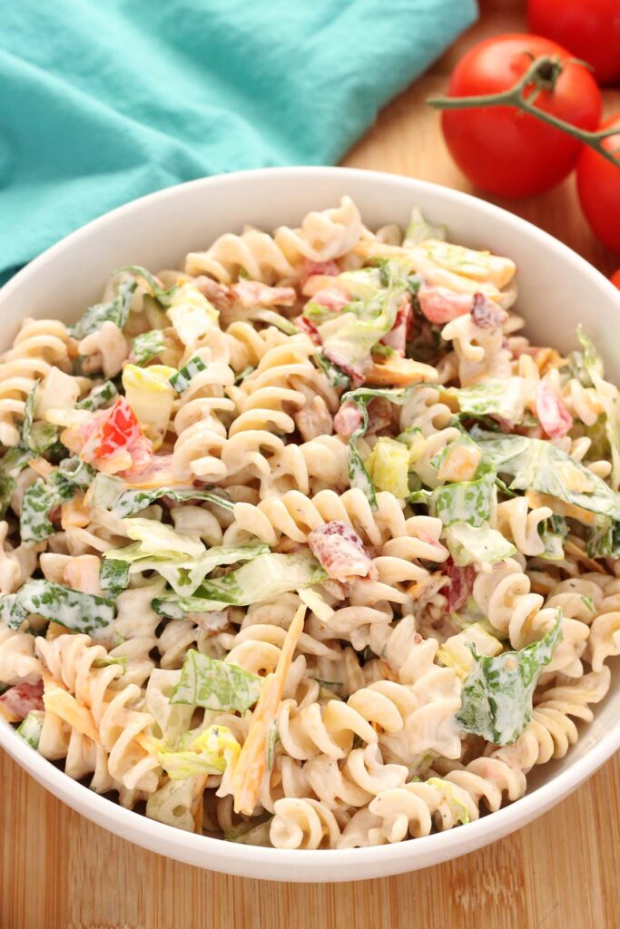 close up image showing a white serving dish filled with a creamy pasta salad that included tomatoes, cheddar cheese, lettuce and bacon
