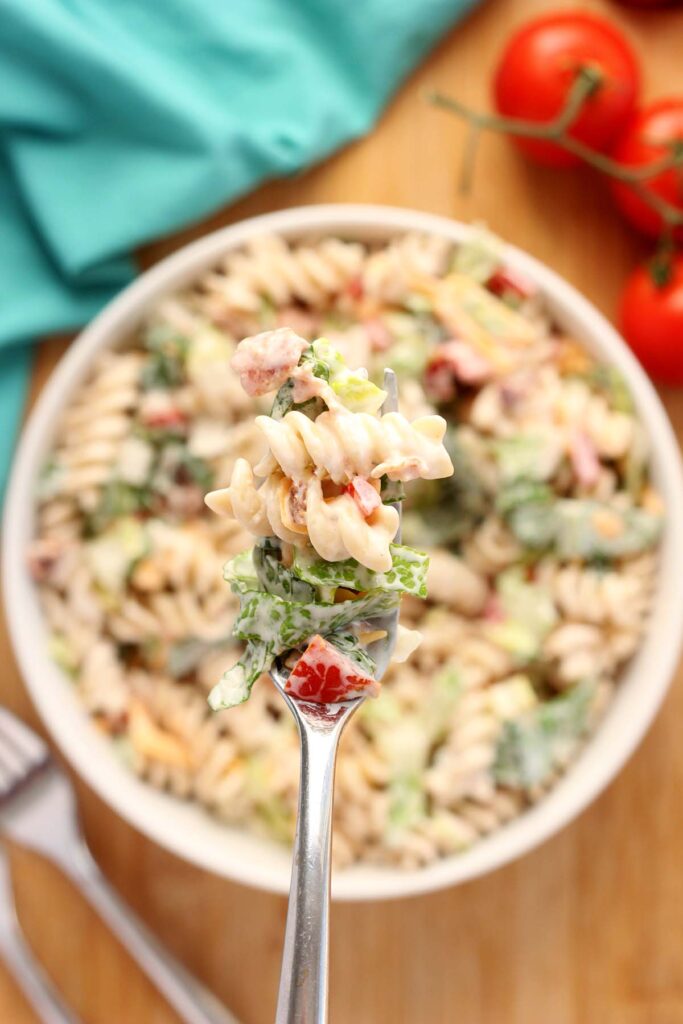 close up image showing a white serving dish filled with a creamy pasta salad that included tomatoes, cheddar cheese, lettuce and bacon with a bite on a fork 