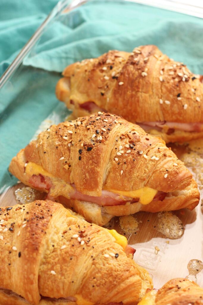 close up image showing a glass dish containing croissant sandwiches filled with ham and cheese and topped with bagel seasoning
