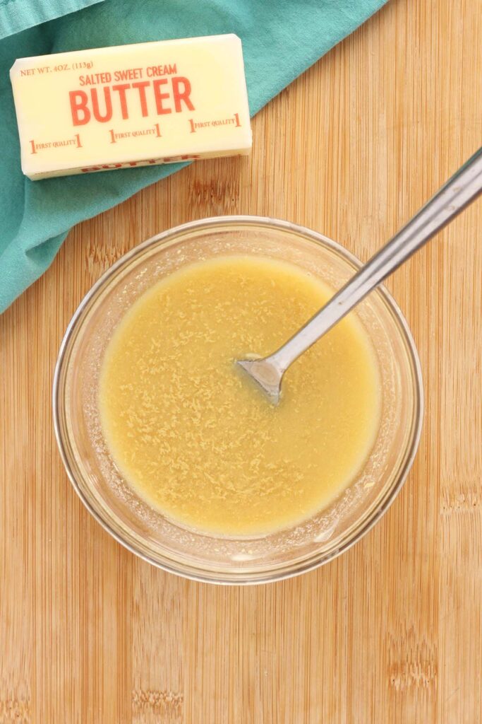 top down image showing a glass bowl filled with melted butter and mustard  whisked together with a fork on a wooden table top