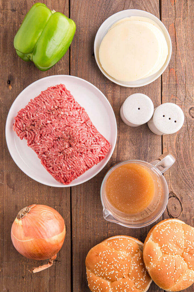 top down iamge showing ground beef, cheese, salt and pepper, broth, onion, pepper and buns sitting on a wooden table top