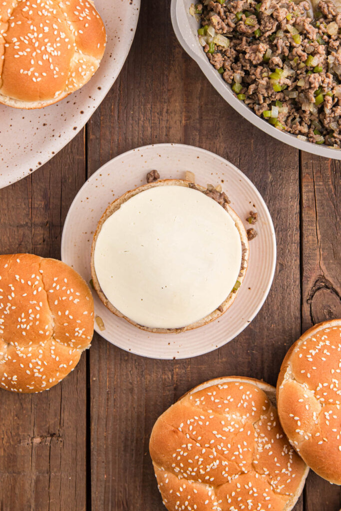 Top down image showing a bun on a white plate topped with a meat mixture and slice of white cheese