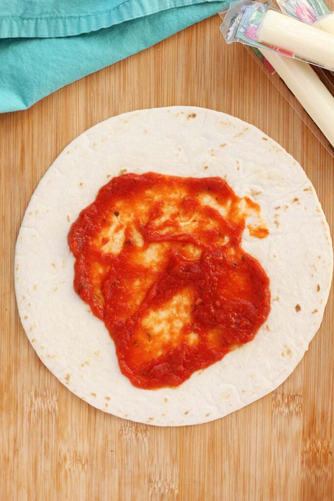 Top down image showing a tortilla sitting on a wooden table top spread with pizza sauce