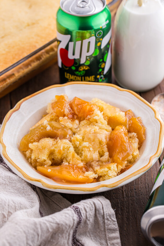 close up image showing a white shallow bowl with a yellow edge filled with peach cobbler. There is a can of 7 up behind the dish