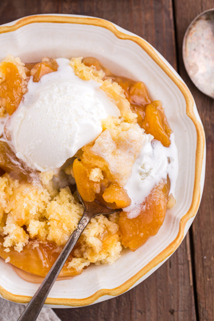 top down image showing a white bowl with a yellow edge filled with peach cobbler and ice cream with a spoon.