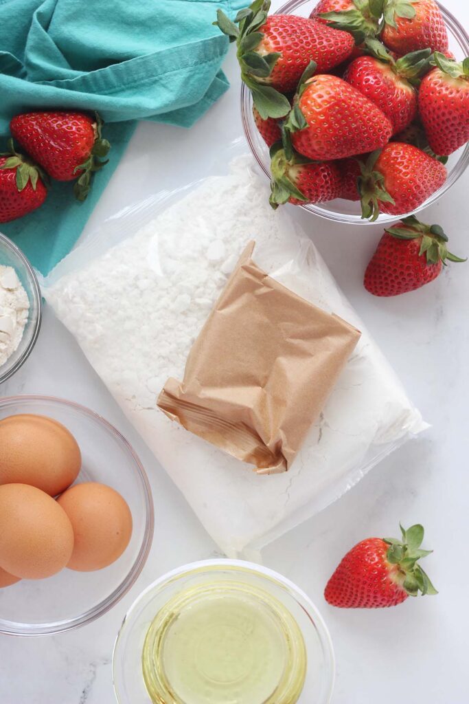 Top down image showing cupcake ingredients ona table top. There is a glass of strawberries, cake mxi, jello mix, eggs, flour and oil