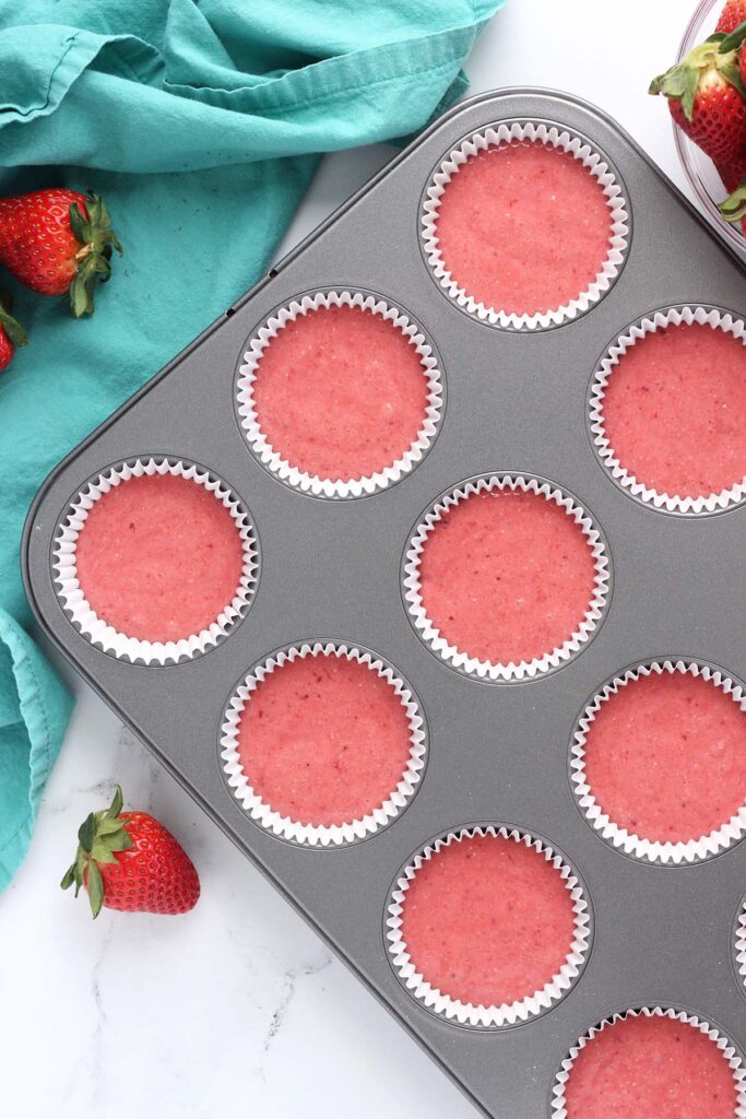 Top down image showing a cupcake tin filled with white liners that are filled with strawberry cake batter. There are fresh strawberries and teal napkins off to the side
