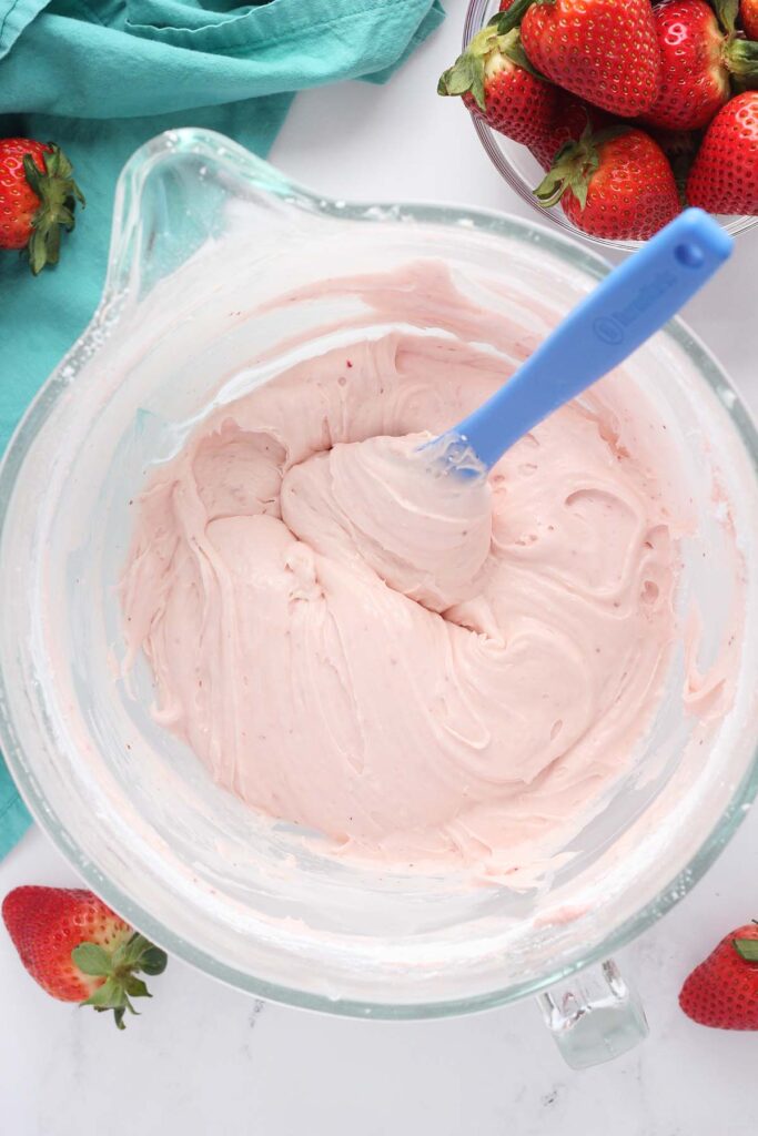 Top down image showing a glass mixing bowl filled with strawberry frosting and a blue spatula. There are fresh strawberries and a teal napkin around the bowl
