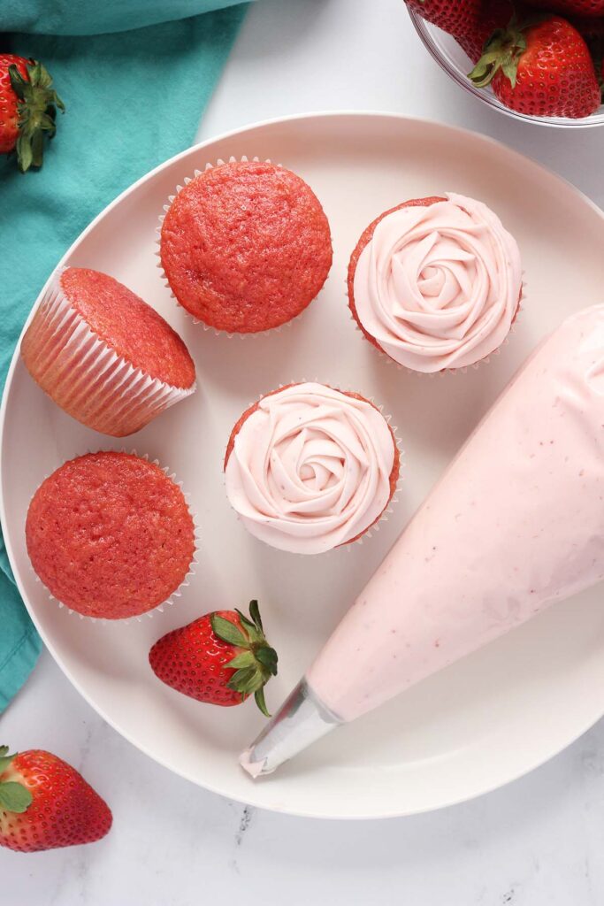 top down image showing three unfosted pink cupcakes and two frosted cupcakes. There is a piping bag filled with frosting with fresh strawberries around