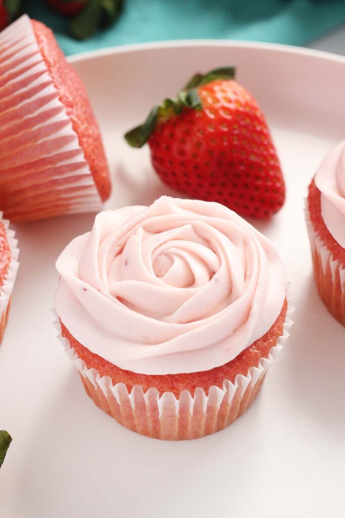 close up image showing a white plate with pink cupcakes. The frosting is piped to look like a rose. there is a strawberry in the background with a teal napkin