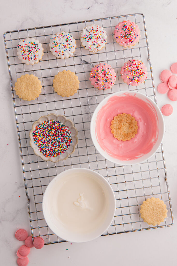 top down image showing cookies being dipped into pink and white chocolate with rainbow sprinkles on top of a wire cookie rack