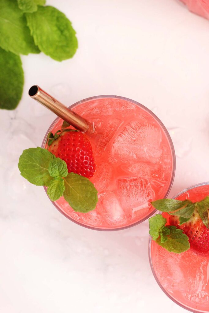 top down image showing a glass filled with ice and a pink soda drink. There is a strawberry and mint inside with a gold straw