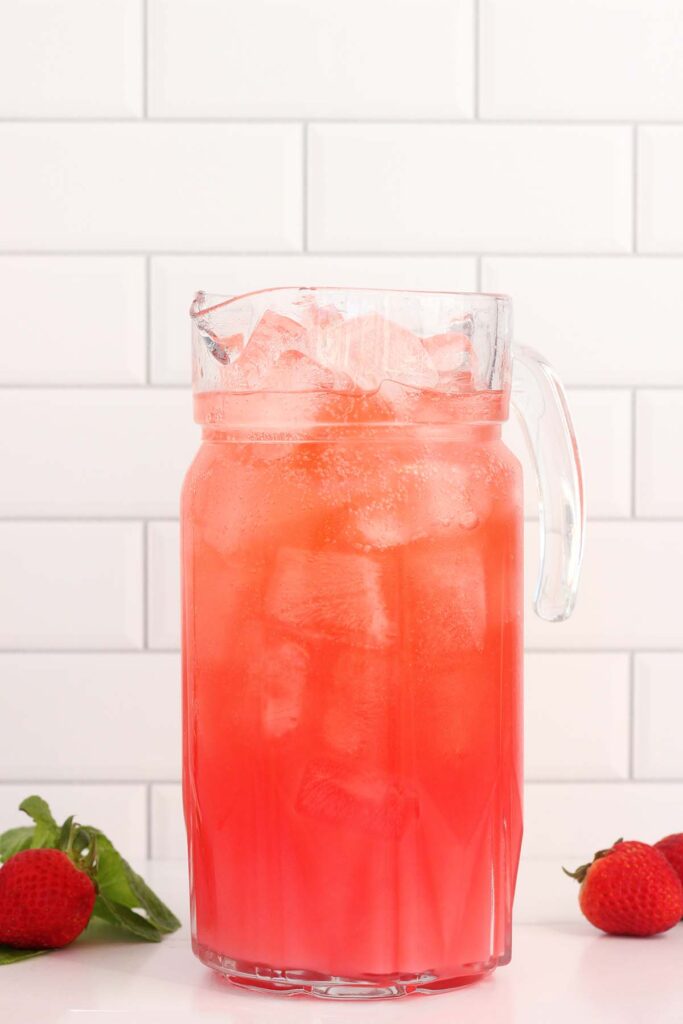 close up image showing a pitcher filled with a red drink and ice with strawberries off to the sides
