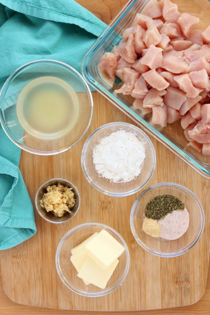 Top down image showing chopped chicken pieces in a bowl, chicken broth, flour, seasonings, garlic, and butter in individual glass bowls with a teal napkin off to the side