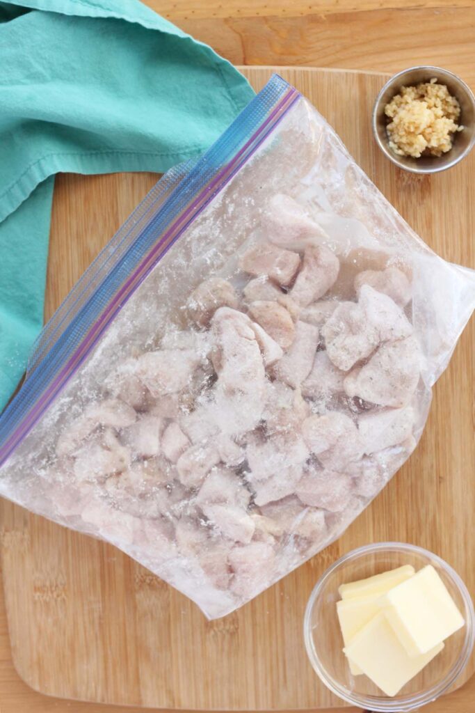 Top down image showing a zip top bag filled with cornstarch covered chicken pieces sitting on a wooden table top. Garlic and butter in small bowls next to it