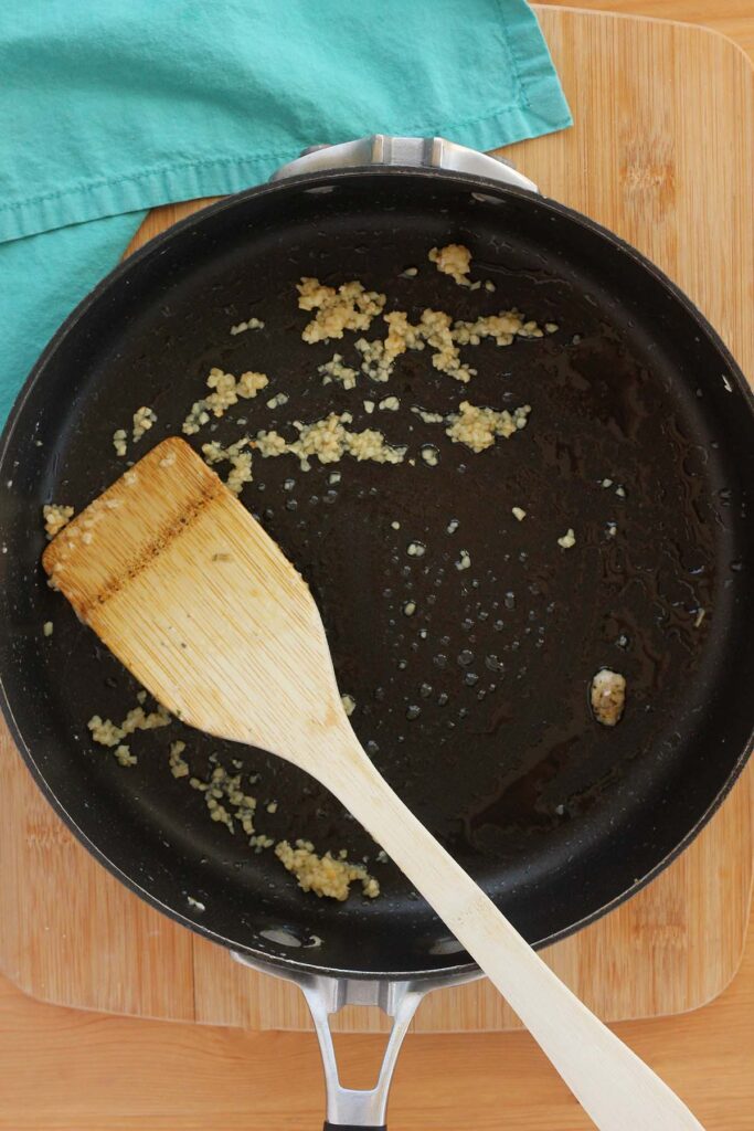 Top down image showing garlic being cooked inside a black skillet with a wooden spoon