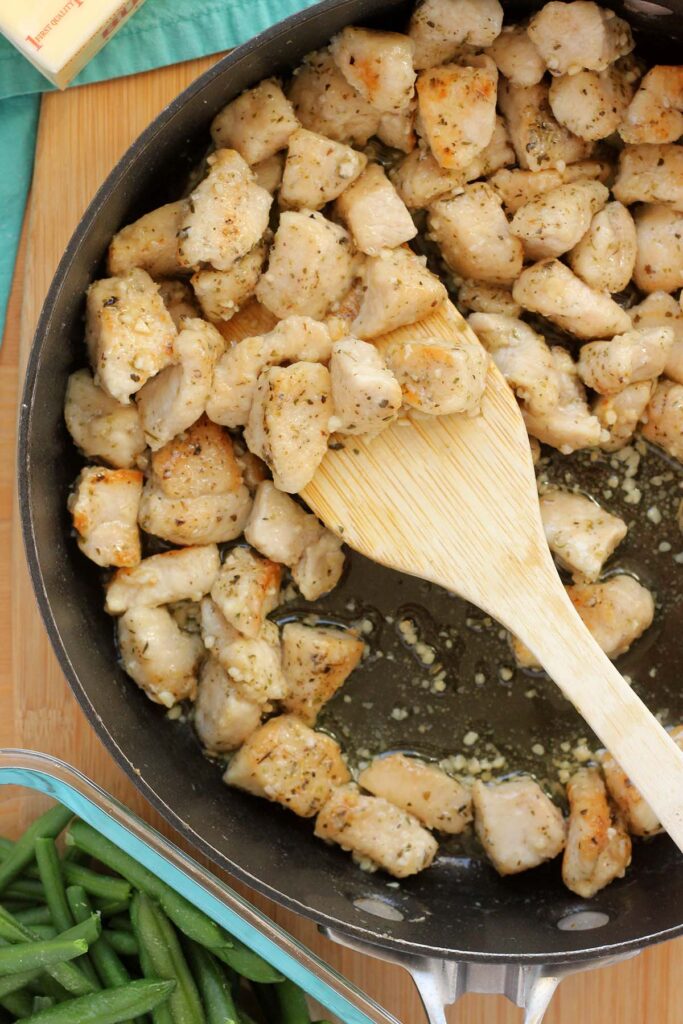 Top down image showing cooked chicken pieces in garlic in a skillet with a wooden spoon