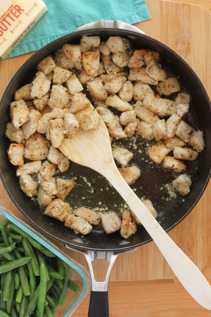 Top down image showing cooked chicken pieces in garlic in a skillet with a wooden spoon