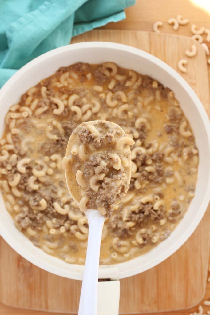 Top down image showing a white skillet filled with a cheesy macaroni pasta with ground beef with a white serving spoon holding a scoop
