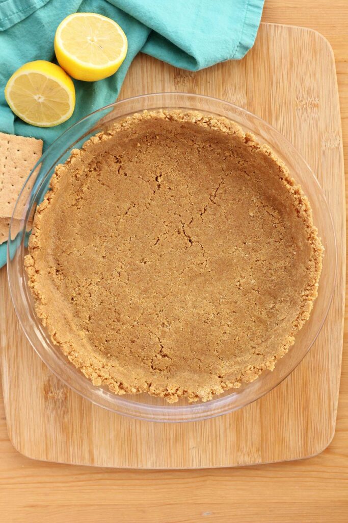 top down image showing a glass mixing bowl filled with crushed graham crackers and melted butter that have been pressed down to form a crust