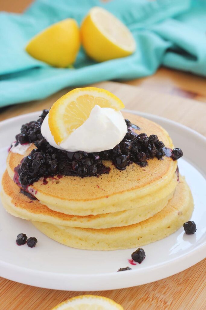close up image showing a stack of 3 pancakes topped with lemon, blueberries, and whipped cream on a white round plate with a lifted lip