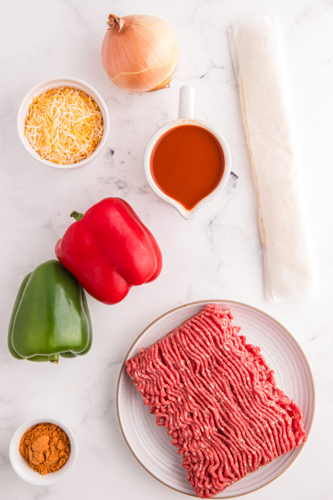 Top down image showing pizza crust, ground beef, onion, cheese, enchilada sauce, seasoning, and bell peppers on a white table top