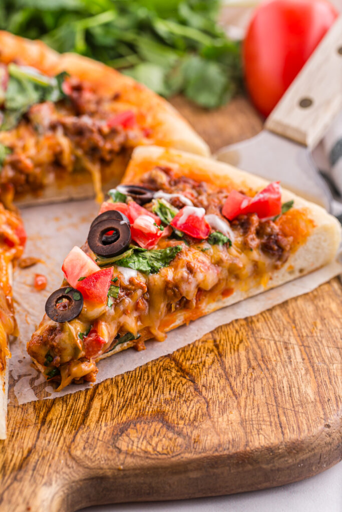 close up on a single slice of pizza that is layered with a beef mixture, cheddar cheese, lettuce, tomato and olives