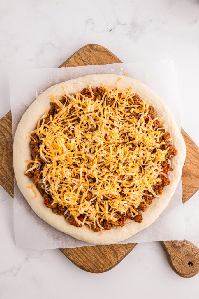 Top down image showing a pizza dough rolled out and shaped on a piece of parchment with a beef topping and sprinkled with cheese