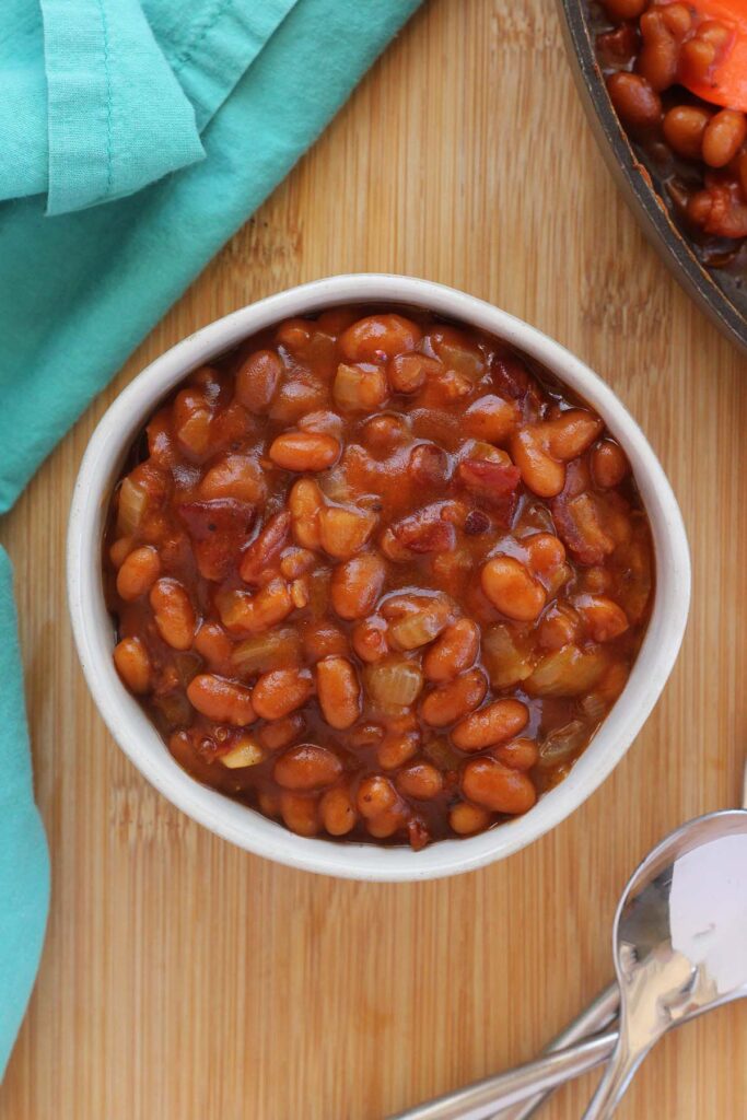 Top down image showing a small white bowl filled with beans. The bowl is on a wooden tabel top with a teal napkin and spoons