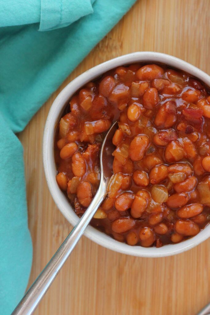 Top down image showing a small white bowl filled with beans. The bowl is on a wooden tabel top with a teal napkin and spoons