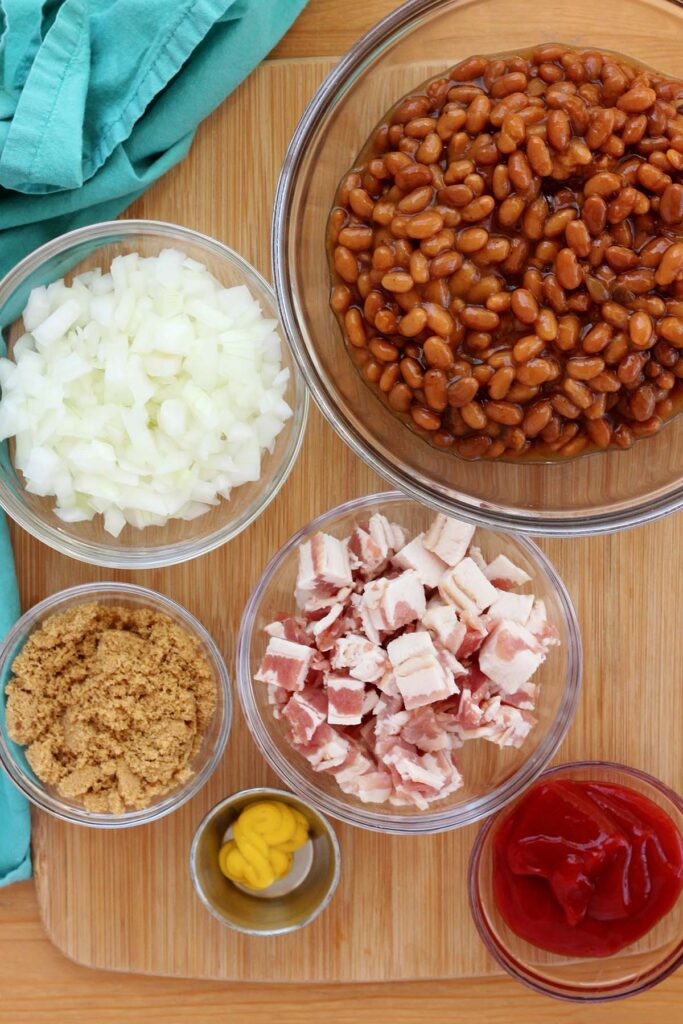 top down image showing a glass bowl filled with beans, a bowl filled with chopped onion, one with chopped bacon, one brown sugar, ketchup and mustard
