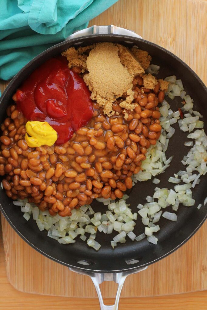 Top down image showing diced cooked onion inside a black skillet with beans, brown sugar, ketchup and mustard