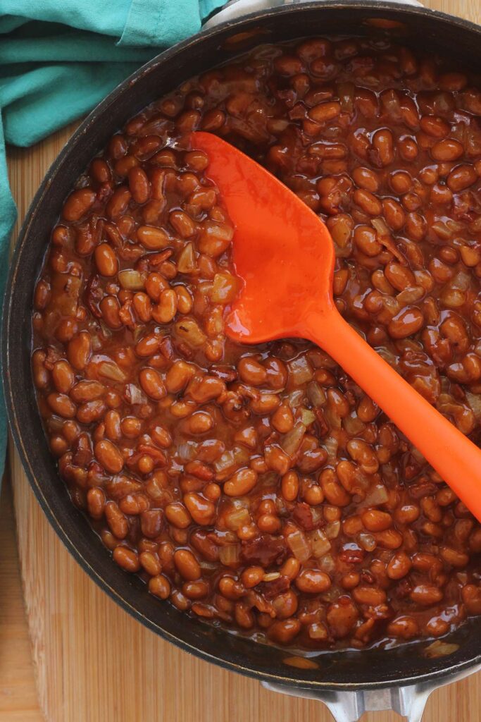 close up image showing a black skillet filled with saucy beans being stirred with an orange silicone spatula