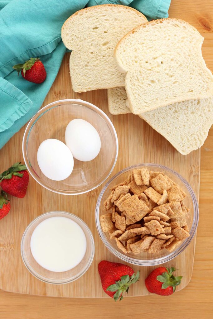 Top down image showing a stack of white bread, cinnamon french toast cereal, two eggs, and milk with strawberries and a teal towel.