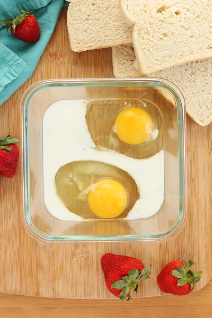 Top down image showing a square bowl on a wooden table top with two eggs and milk inside. Strawberries, bread, and a teal towel off to the side