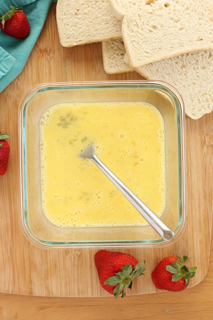 Top down image showing a square bowl on a wooden table top with two eggs and milk whisked inside. Strawberries, bread, and a teal towel off to the side
